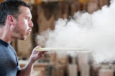Caucasian man factory worker blowing sanding dust off a wooden piece in ...
