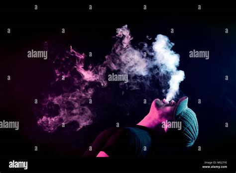 A Young Man Exhales A Cloud Of Colored Smoke Of Red Color On A Black