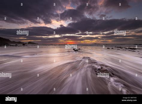Waves Flow Over The Sand And Rocks At Sunset At Welcombe Mouth Beach In