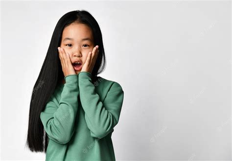 Шокированная удивленная азиатская девочка подросток в зеленой одежде трогает щеки Премиум Фото