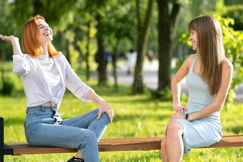 Two Young Girls Friends Sitting On A Bench In Summer Park Chatting