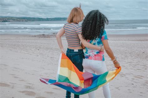 joven pareja de lesbianas moviendo la bandera del orgullo gay en una playa de arena mientras