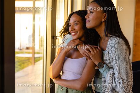 Happy Biracial Lesbian Couple Embracing And Looking Out Window At Home Lifestyle Relationship