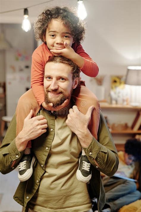Happy Young Man Looking At Camera While Holding His Son On Shoulders