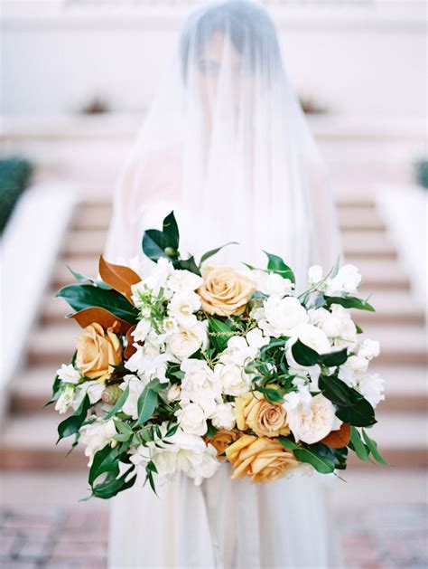A Bride Holding A Bouquet Of White And Yellow Flowers In Front Of A