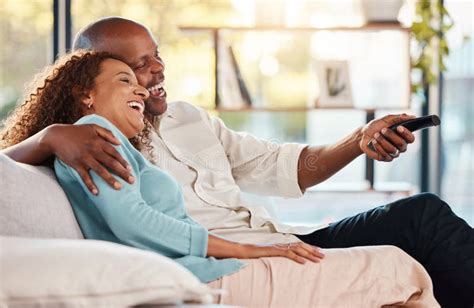 Couple Watching Tv And Laughing On Sofa In Home Living Room Bonding And Hug Interracial