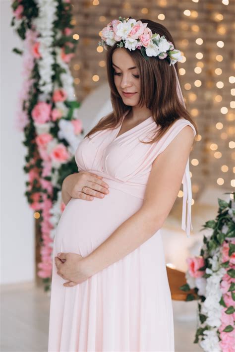 Pregnant woman with drown hair, in pink powder dress, embracing belly. Charming brunette pregnant woman in pink dress poses in ...