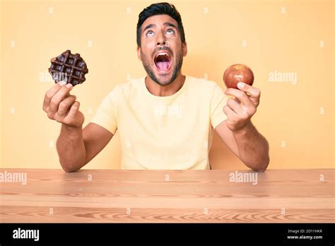 Young Hispanic Man Eating Breakfast Holding Chocolate Waflle And Apple