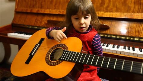 Little Baby Girl Plays Guitar In Front Of Piano With A Red Heart As