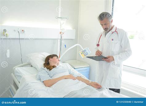Doctor With Clipboard Standing Near Patient Lying On Bed Stock Photo