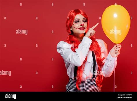 happy woman with bright hair and clown makeup pointing at yellow balloon isolated on red stock