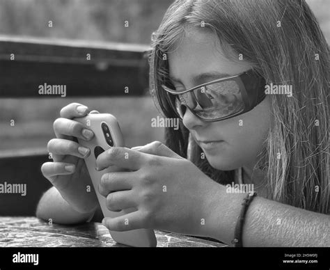 Grayscale Shot Of An Italian Girl Sitting Outdoors Wearing Sunglasses Using Her Smartphone Stock