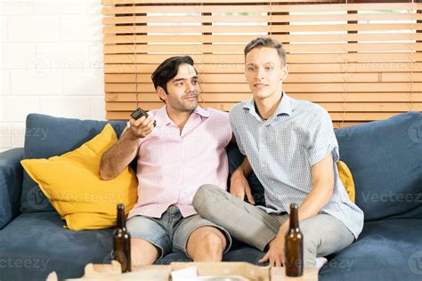 Dos Gay Hombres En Casual Trajes Sentado En Sofá Abrazando Y Acecho Televisión Con Cerveza Y