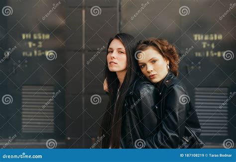 Closeup Portrait Of A Pair Of Girls In Leather Jackets Posing At The Camera Against A Dark Wall