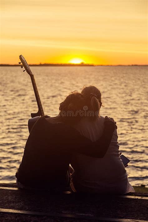 Lesbian Couple Embracing While Watching The Sunset Over The Lagoon Sitting On A Bench Stock