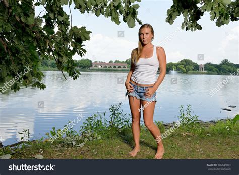 Sexy Blonde Woman In White Tank Top And Denim Cut Off Shorts Standing Barefoot Next To Lake