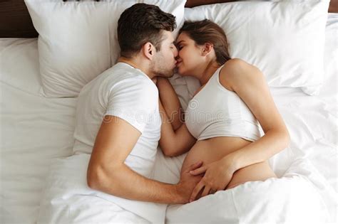 Happy Pregnant Woman Lies In Bed With Her Husband Kissing Stock Photo