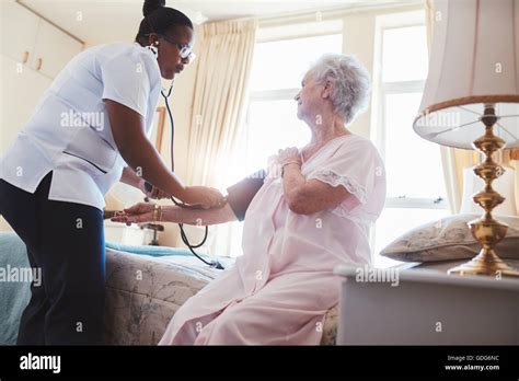 Female Nurse Checking Blood Pressure Of A Senior Woman Sitting On Bed