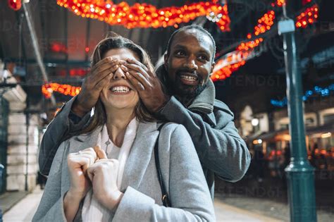 Man Covering Eyes Of Girlfriend At Illuminated Christmas Market Stock Photo