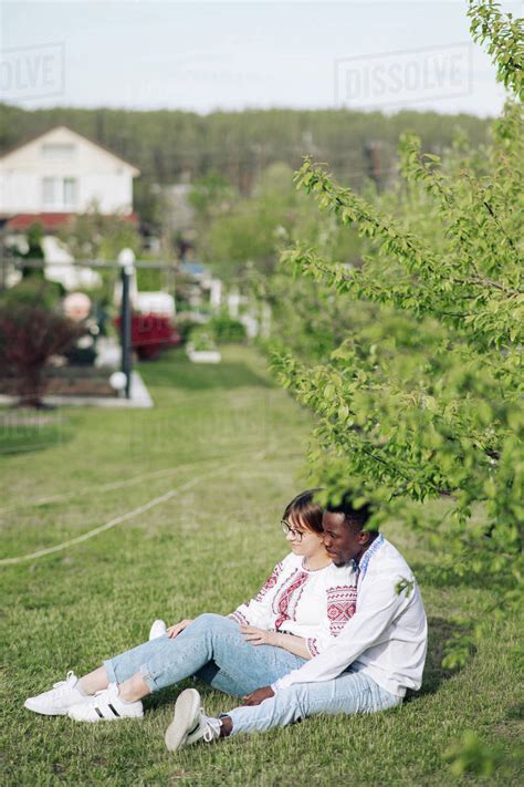 Interracial Couple Sits On Grass In Spring Garden Dressed In Ukrainian Traditional Ethnic