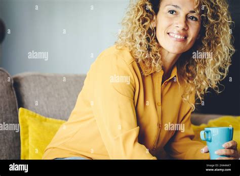 happy woman smile and look outside at home sitting on the sofa drinking from a mug yellow shirt