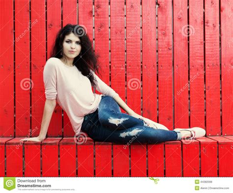 Beautiful Girl With Long Hair Brunette In Jeans Sits Near Wall Of Red
