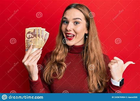 Young Blonde Woman Holding 5000 Hungarian Forint Banknotes Pointing Thumb Up To The Side Smiling
