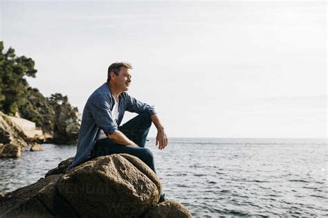 Thoughtful Mature Man Sitting On Rock Against Clear Sky Stock Photo
