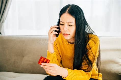 An Ill Asian Woman Sits On A Couch Taking Medicine Consulting Doctor Via Phone Stock Image