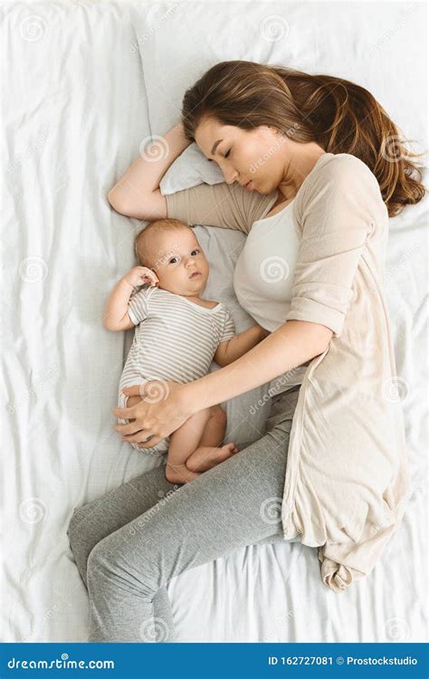 Exhausted Mother Sleeping And Embracing Her Awakened Newborn Baby Stock