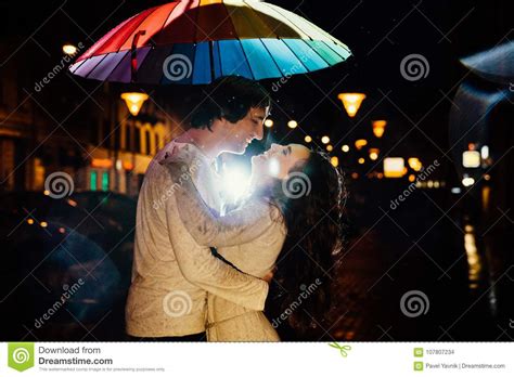 Young Couple Under An Umbrella Kisses At Night On A City Street Stock