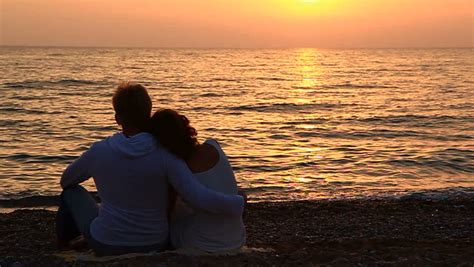 Couple In Love Sitting On The Rocks Near The Sea They Look At The Water And The Rising Sun The