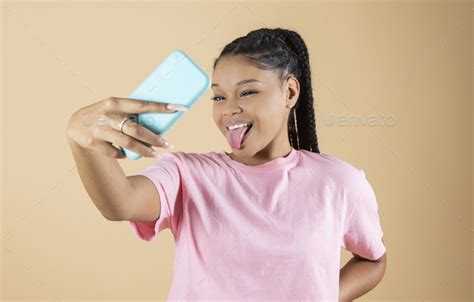 Mixed Race Woman Takes A Selfie With Her Smartphone Sticking Out Her Tongue On Yellow Background