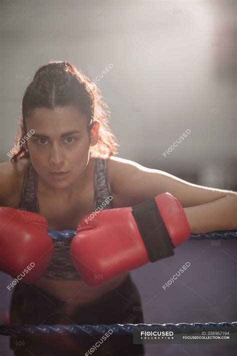 Portrait Of Female Boxer In Gloves Leaning On Boxing Ring Rope At