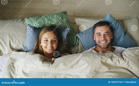 Top View Of Smiling Couple Having Fun In Bed Hiding Under Blanket And Looking Into Camera Stock