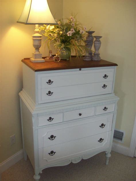 A White Dresser With Flowers On Top And A Lamp In The Corner Next To It