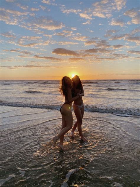 Two Girls Are Standing In The Water At The Beach As The Sun Sets Behind Them