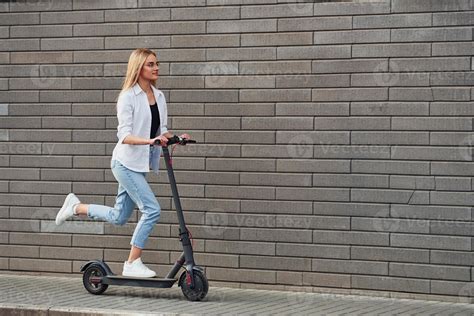 Side View Of Beautiful Blonde In Casual Clothes That Riding Electric Schooter Outdoors At Sunny