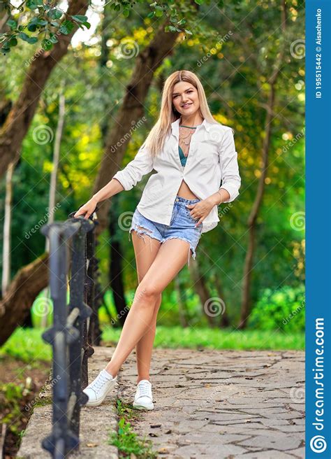 Relaxed Girl In Jeans Shorts And White Shirt On A Bridge In Summer Park