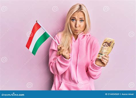 Young Blonde Woman Holding Hungary Flag And Forint Banknotes Puffing Cheeks With Funny Face