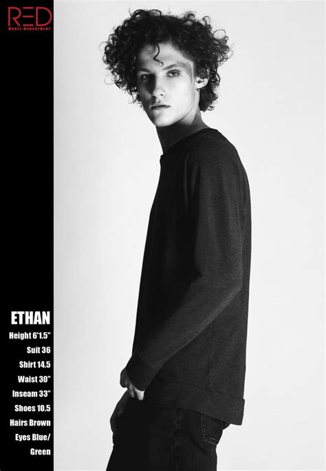 A Man With Curly Hair Standing In Front Of A Black And White Background Wearing A Sweater