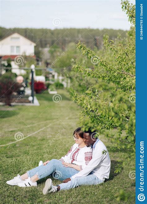 Interracial Couple Sits On Grass In Spring Garden Dressed In Ukrainian Embroidered Shirts Stock