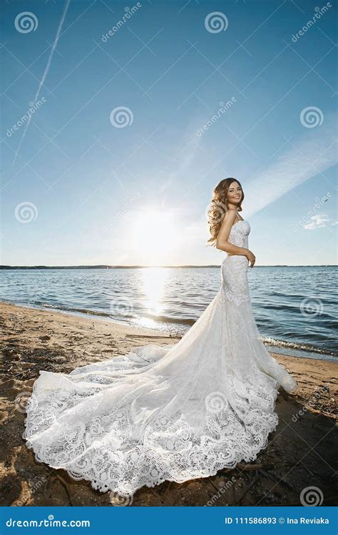 Young Brunette Bride Beautiful Model Girl In White Lace Dress Is Standing On Coast At Sunset