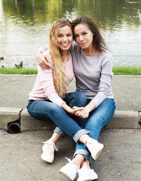 Mom And Her Adult Daughter Are Sitting On The Road Near The Lake Happy
