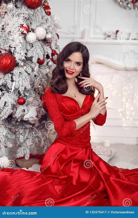 Christmas Portrait Of Happy Smiling Girl In Red Dress Sitting By Snowy Xmas Tree Brunette With