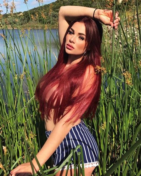 A Woman With Long Red Hair Sitting In Tall Grass Next To The Water And
