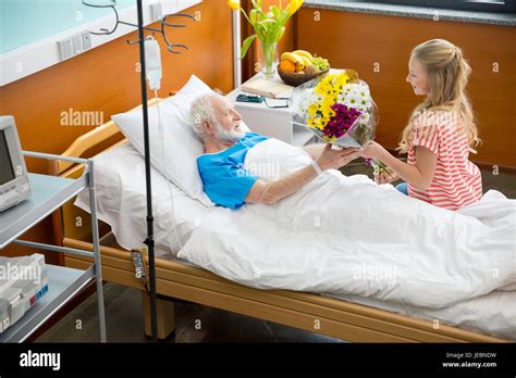 Smiling Girl Giving Flowers To Sick Grandfather Lying In Hospital Bed