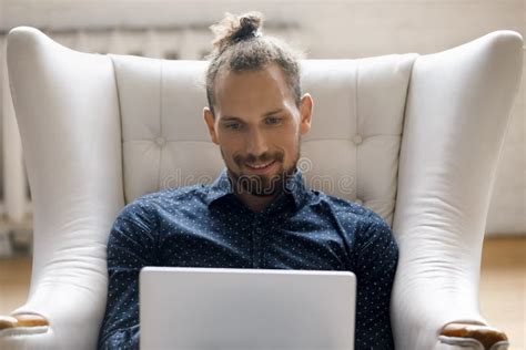 Happy Millennial Hipster Man Using Computer Sitting In Armchair Stock