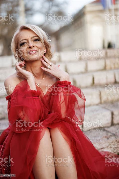 Beautiful Mature Woman In Red Dress With Bouquet Of Red Roses Sitting On Outdoor Staircase Stock