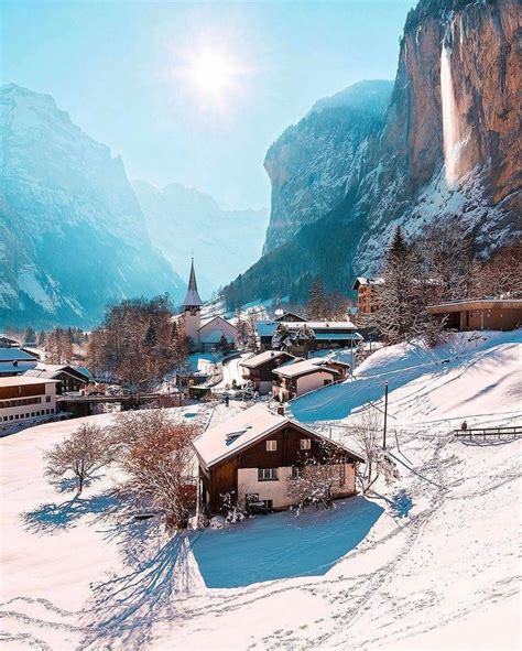 A Snow Covered Village With Mountains In The Background And Sun Shining